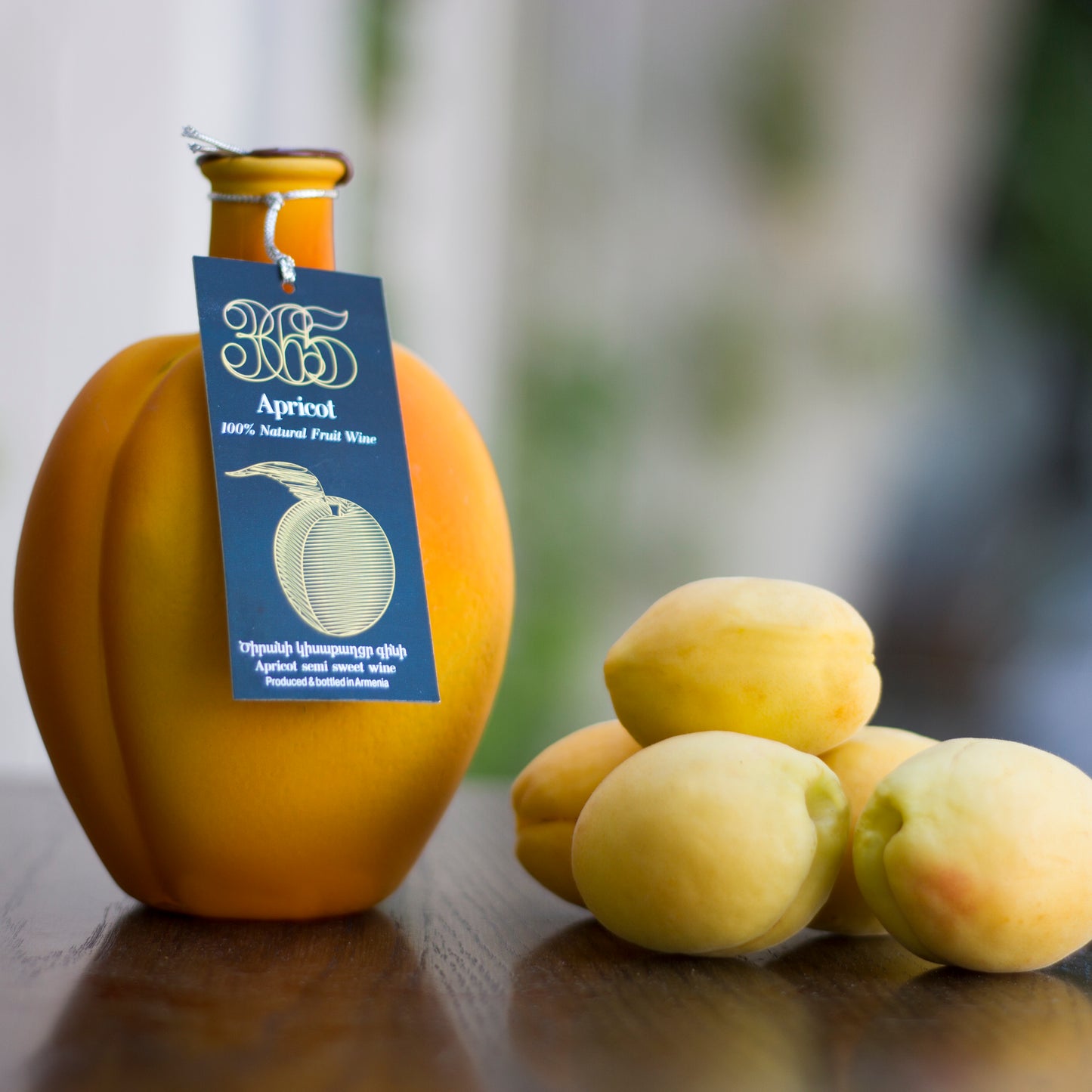 Apricot-shaped bottle of 365 Apricot Semi-Sweet Wine with product tag, placed beside fresh apricots on a wooden surface.