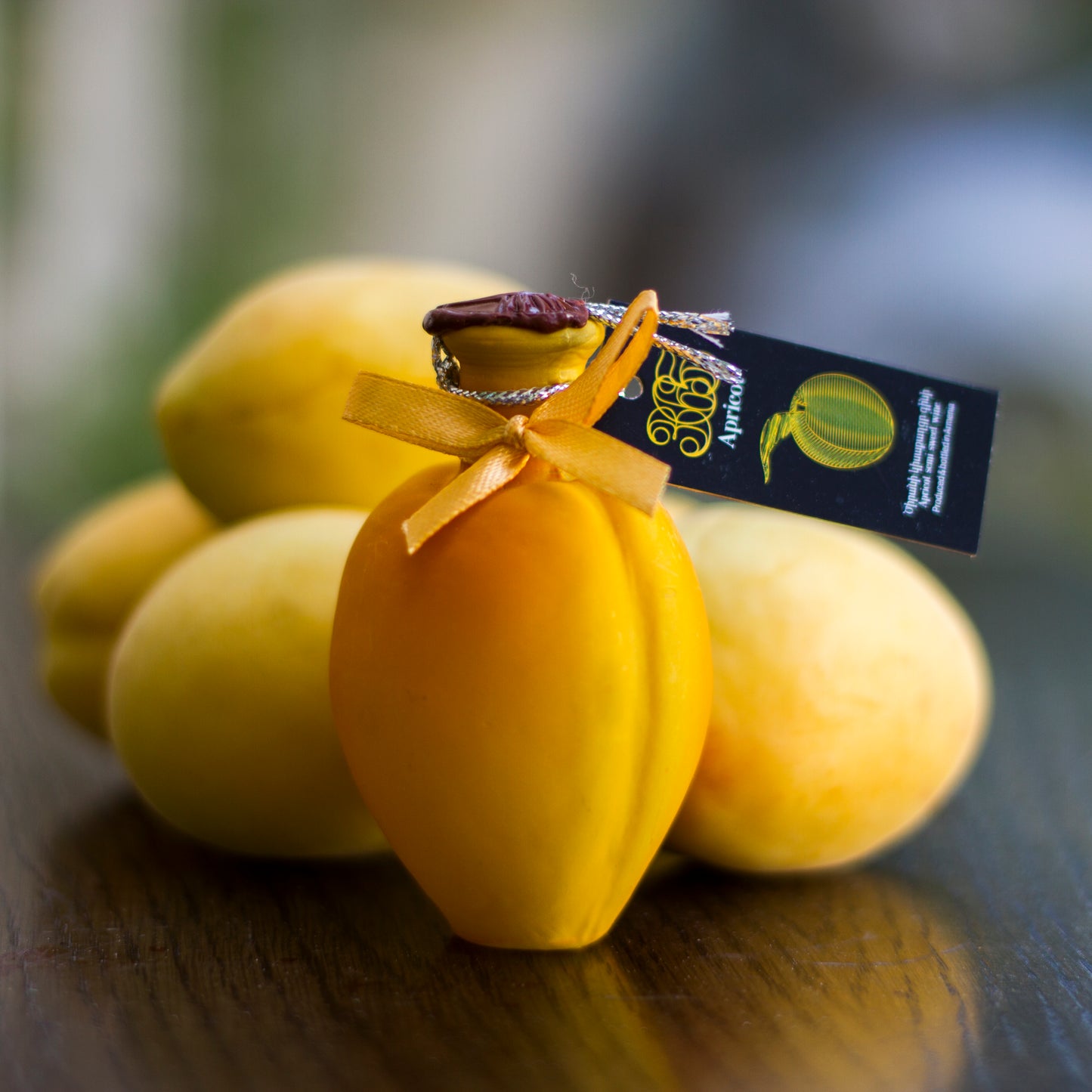 Mini apricot-shaped souvenir bottle of 365 Apricot Semi-Sweet Wine with a golden ribbon and product tag, surrounded by fresh apricots.