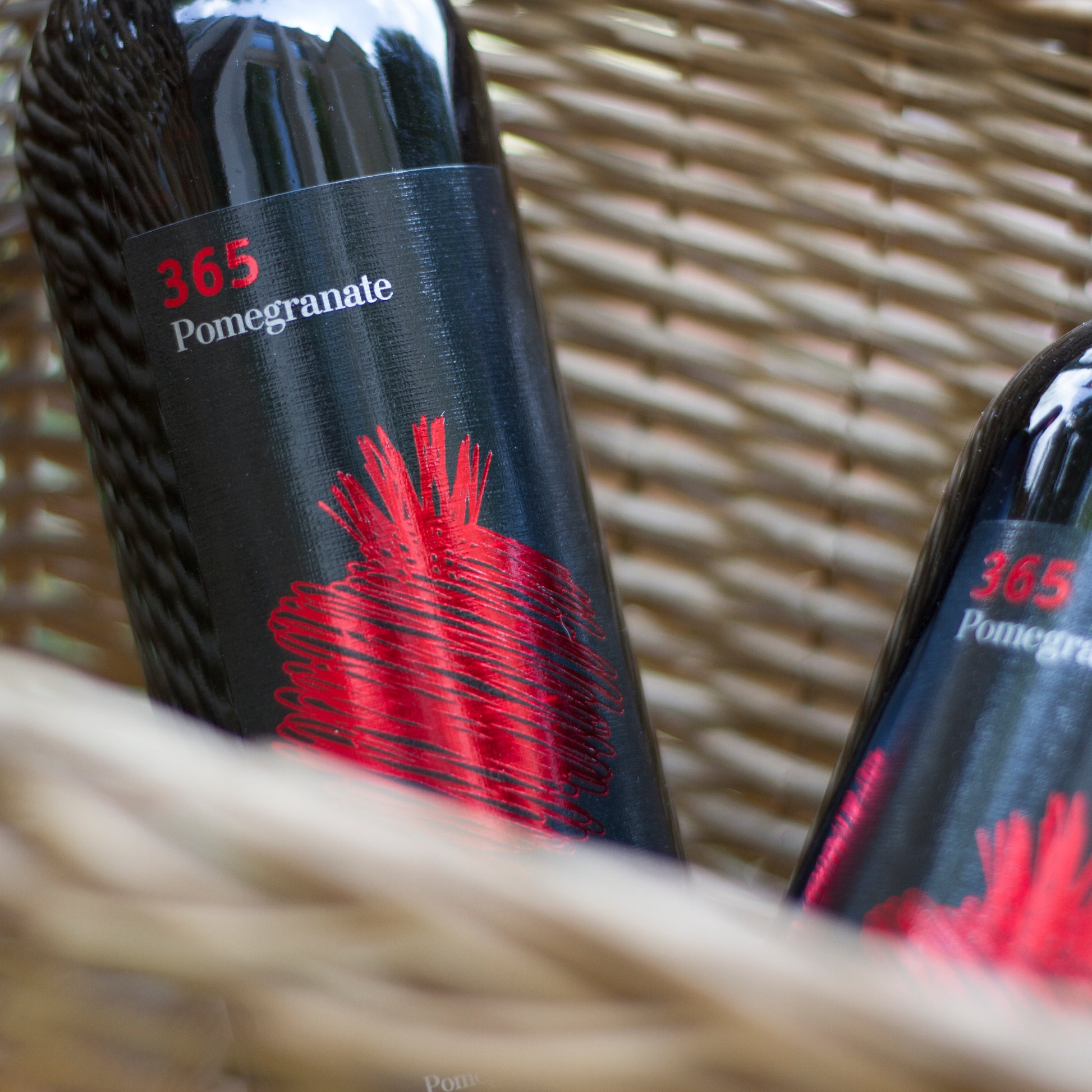 Two bottles of 365 Pomegranate semi-sweet fruit wine resting in a wicker basket with bold red label artwork.