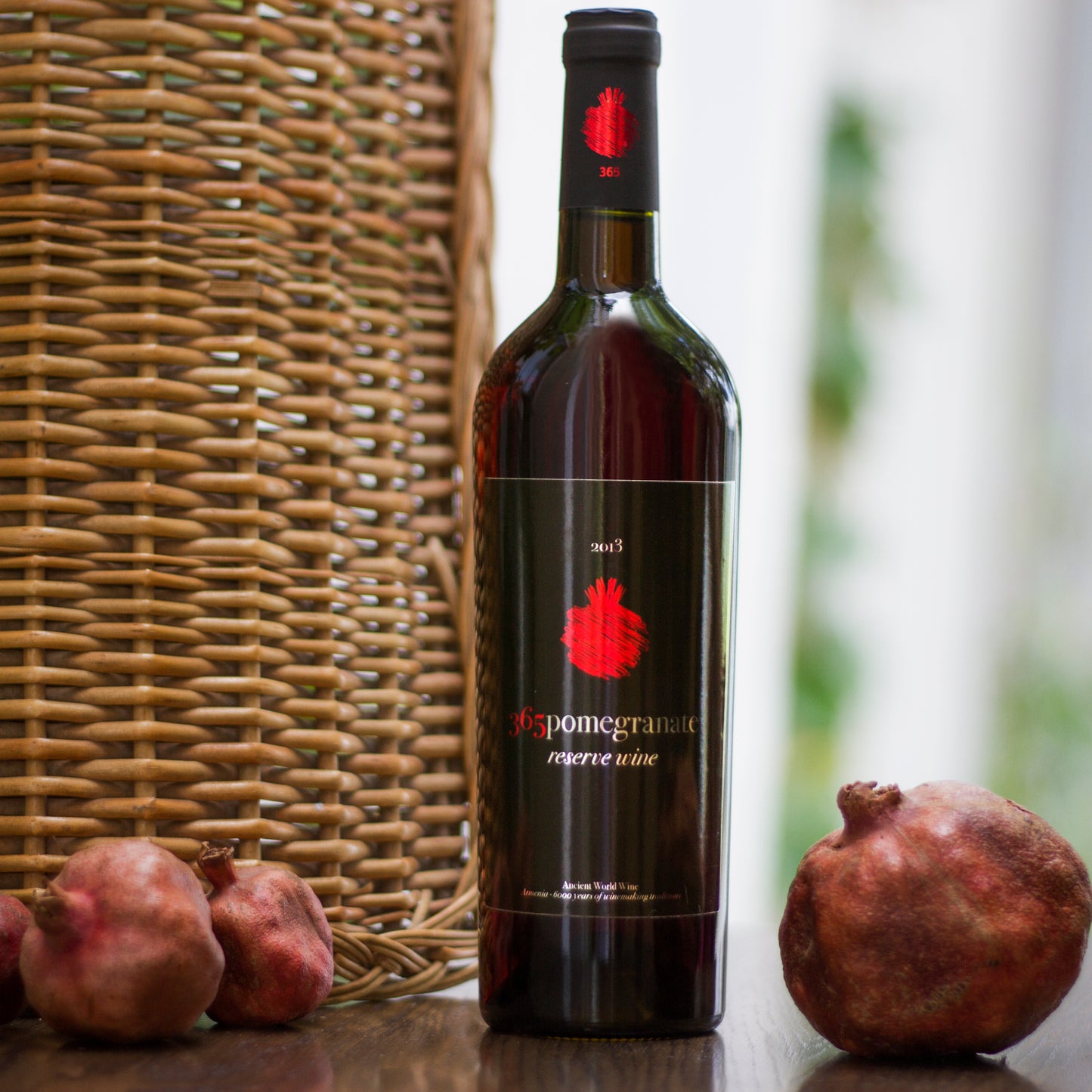 365 Pomegranate Reserve Wine bottle standing beside whole pomegranates with wicker background.