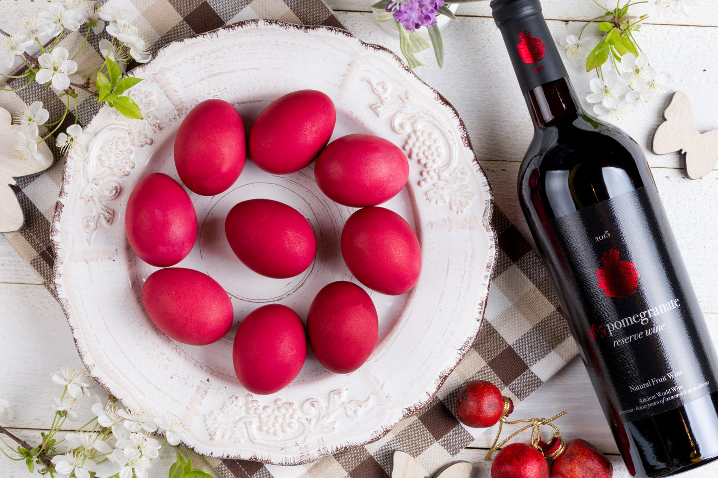365 Pomegranate Reserve Wine bottle with red Easter eggs on a rustic plate and floral spring decor.