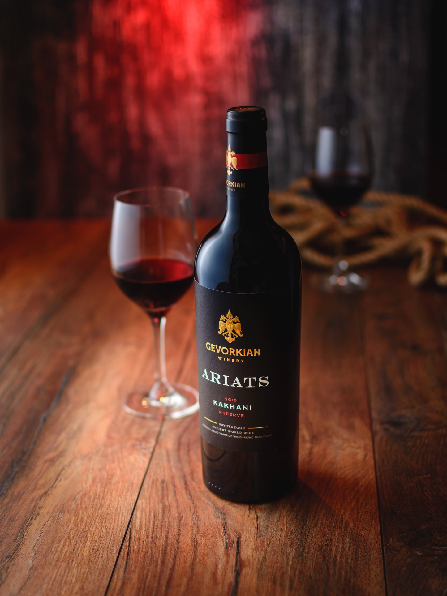 Gevorkian Ariats Kakhani 2019 Reserve red wine bottle with a glass of red wine on a wooden table and a warm red background