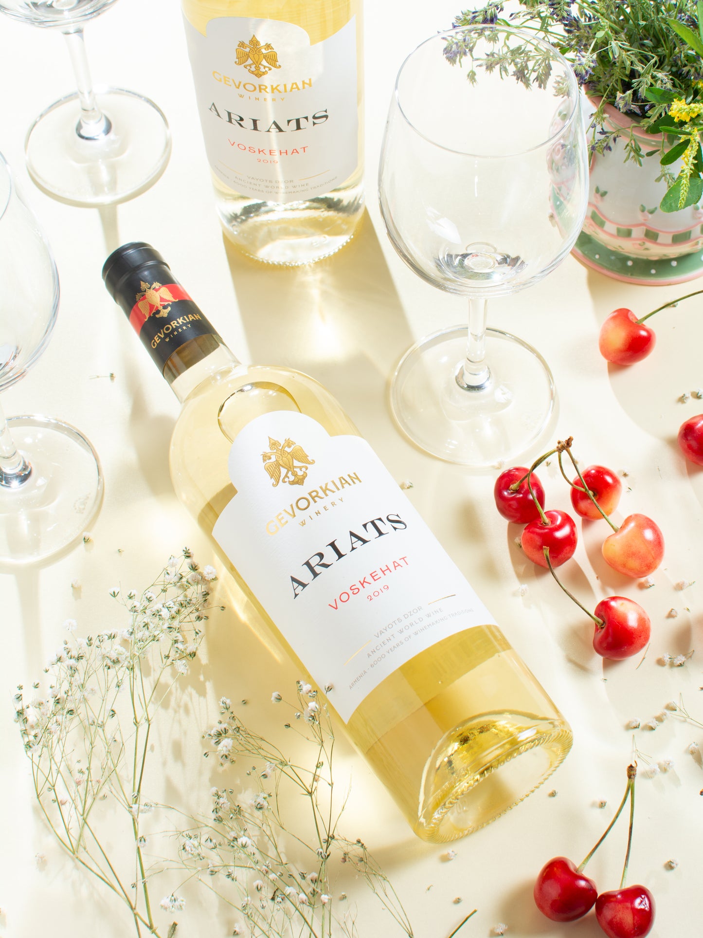Gevorkian Winery Ariats Voskehat 2019 white wine bottle with wine glasses, cherries, and flowers on a light background