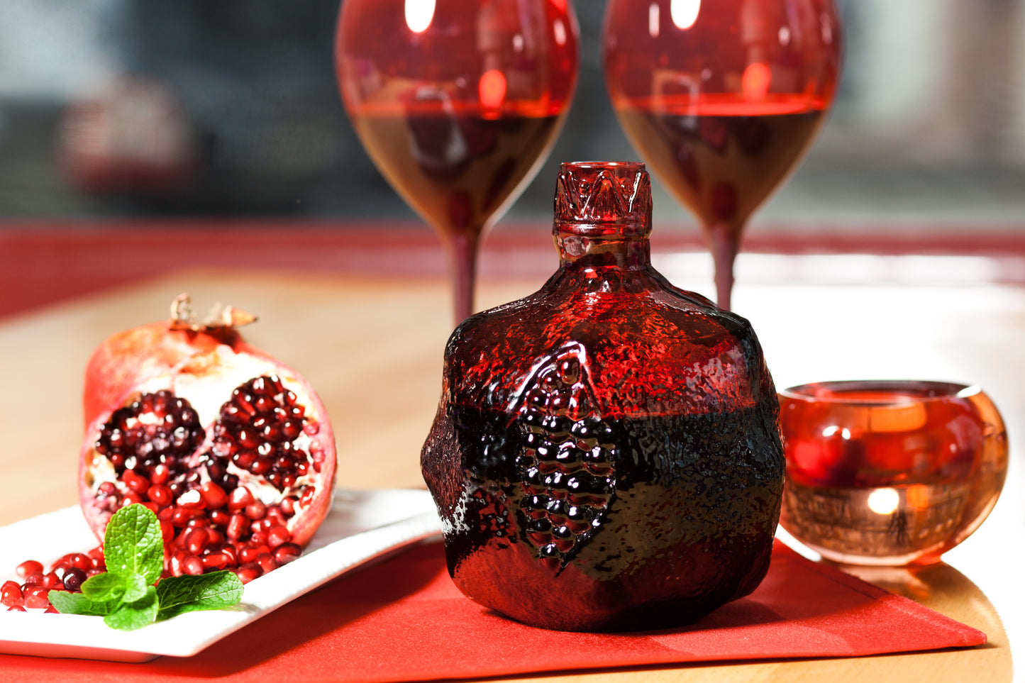 elegant setting featuring the bottle alongside a cut pomegranate, red wine glasses, and mint garnish.