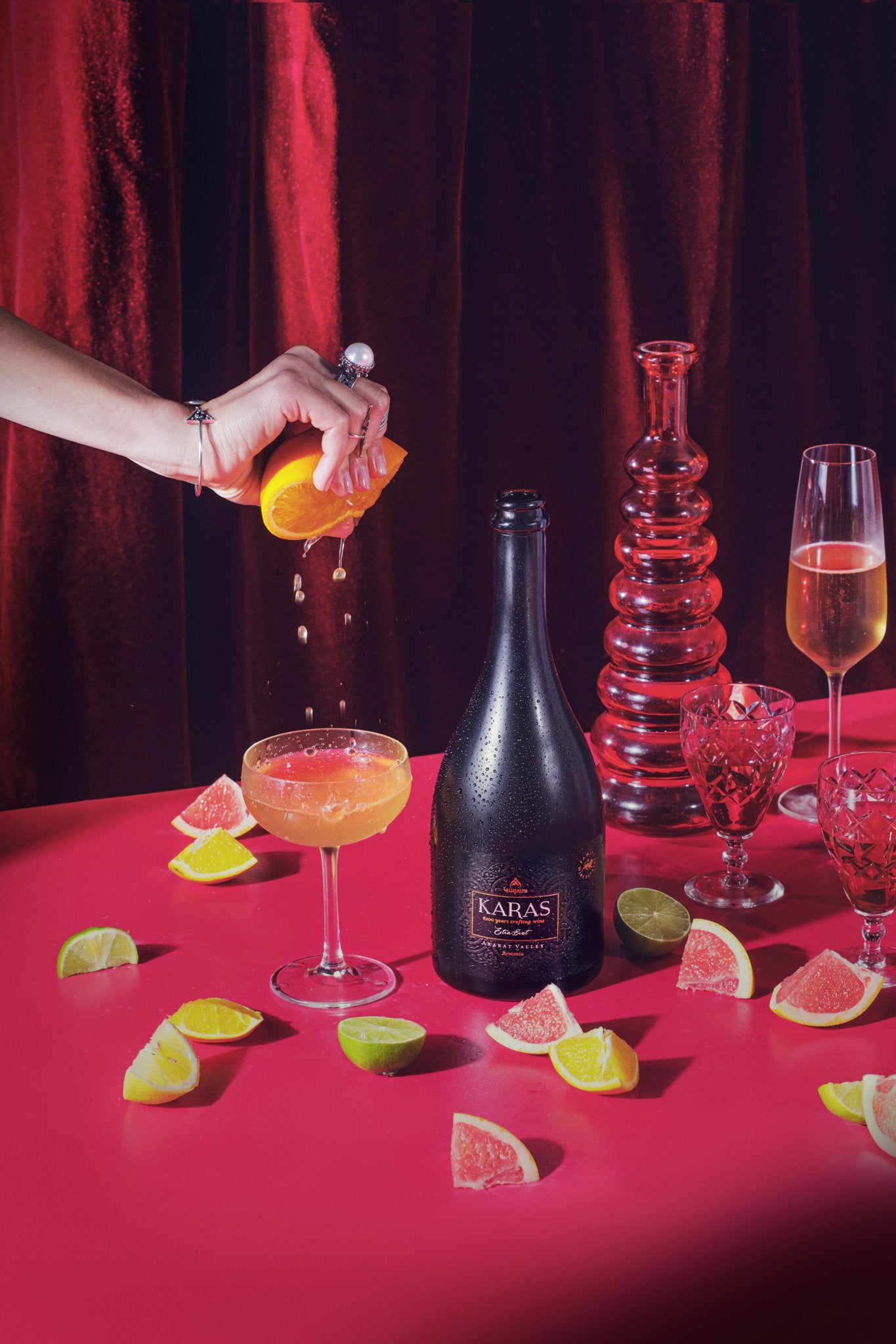 Karas Sparkling Extra Brut bottle on a red table with citrus slices, cocktails, and a hand squeezing orange juice.