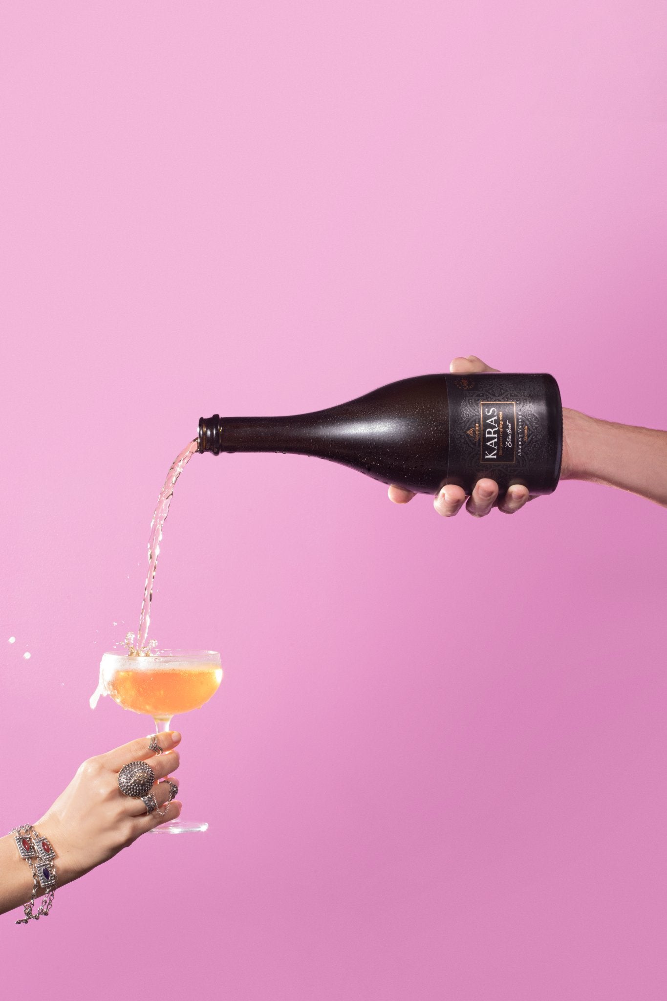 Karas Sparkling Extra Brut being poured into a coupe glass against a pink background, hands in frame.