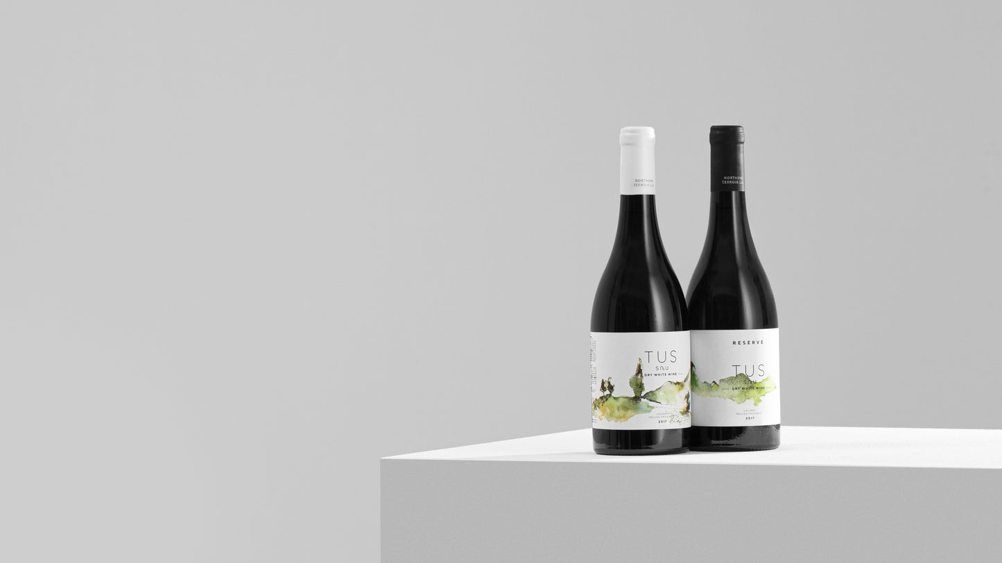 Two bottles of TUS Dry White Wine and Reserve side by side on a white pedestal, photographed in soft black and white studio lighting.