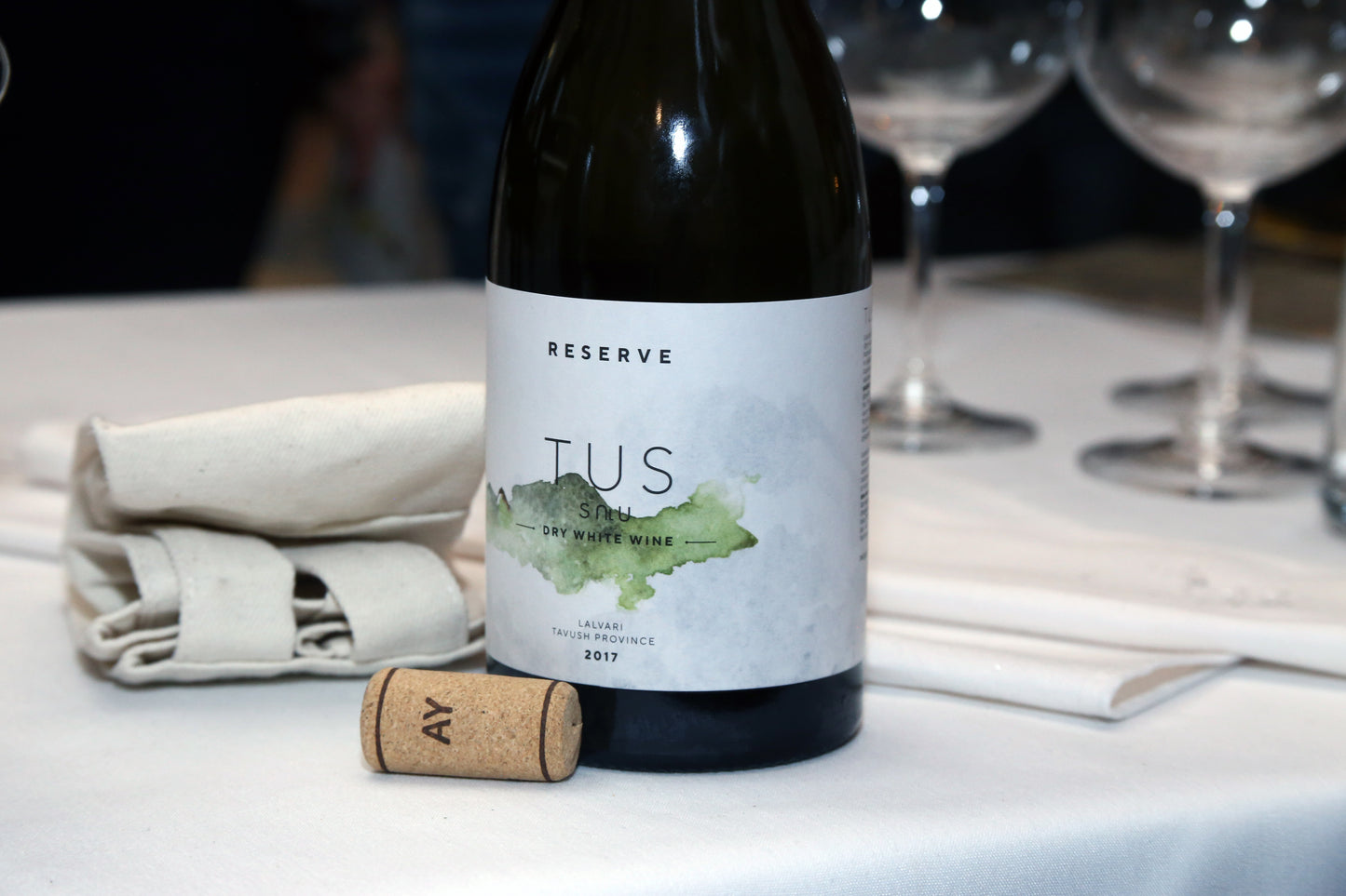 TUS Reserve Dry White Wine 2017 bottle placed on a white tablecloth next to a cork stamped “AY,” with a folded napkin and wine glass in the background.