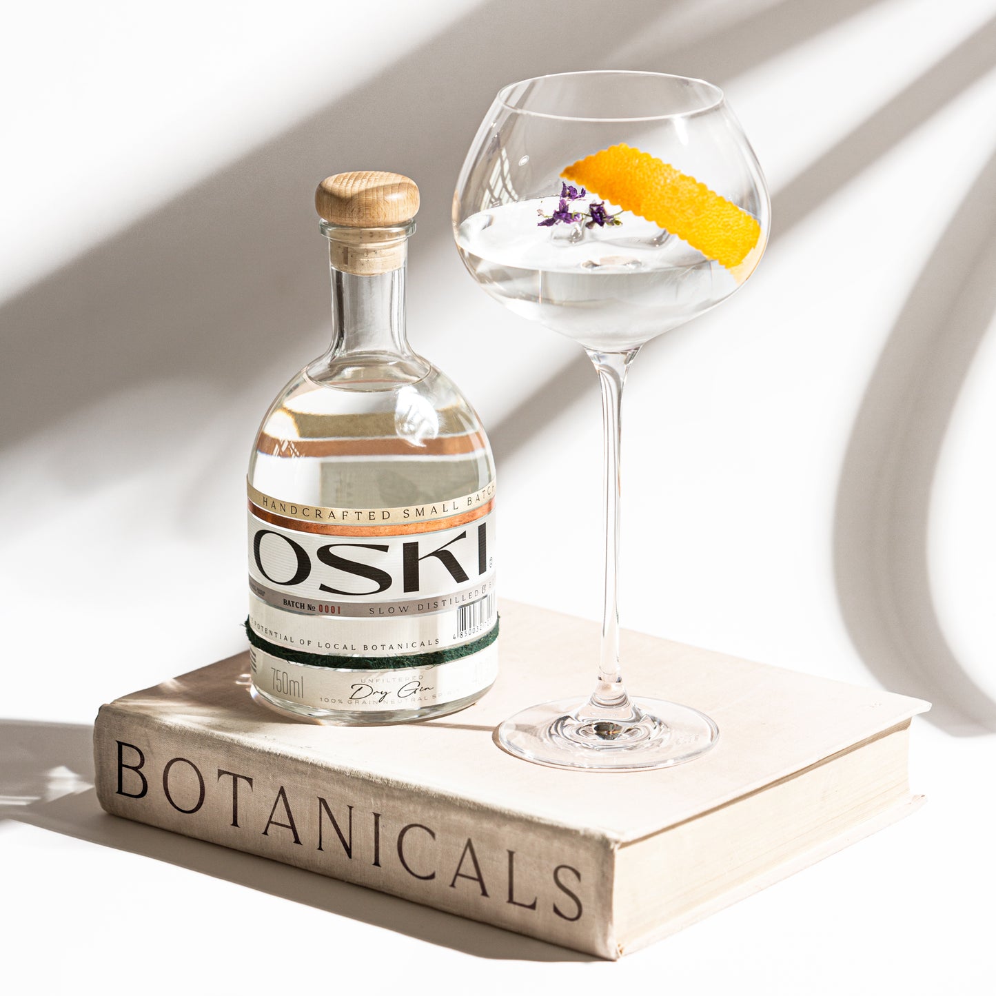 OSKI Gin bottle on top of a "Botanicals" book, paired with a cocktail in a stemmed glass garnished with orange peel and edible flowers.