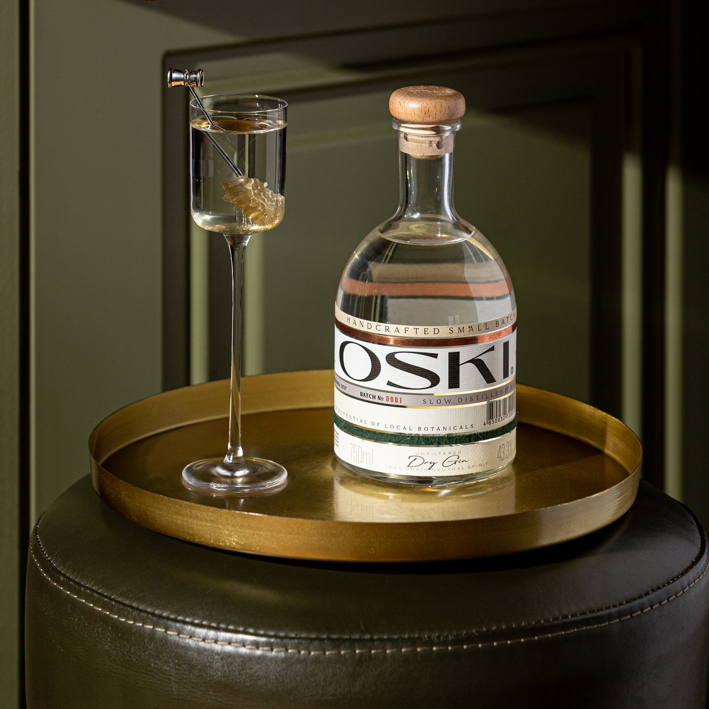 OSKI Gin bottle on a gold tray with a tall glass of gin garnished with a honey stirrer, styled in warm, elegant lighting.