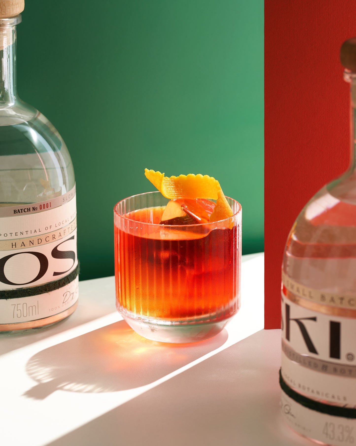 Glass of red cocktail garnished with orange peel, flanked by two OSKI Gin bottles against a bold green and red backdrop.