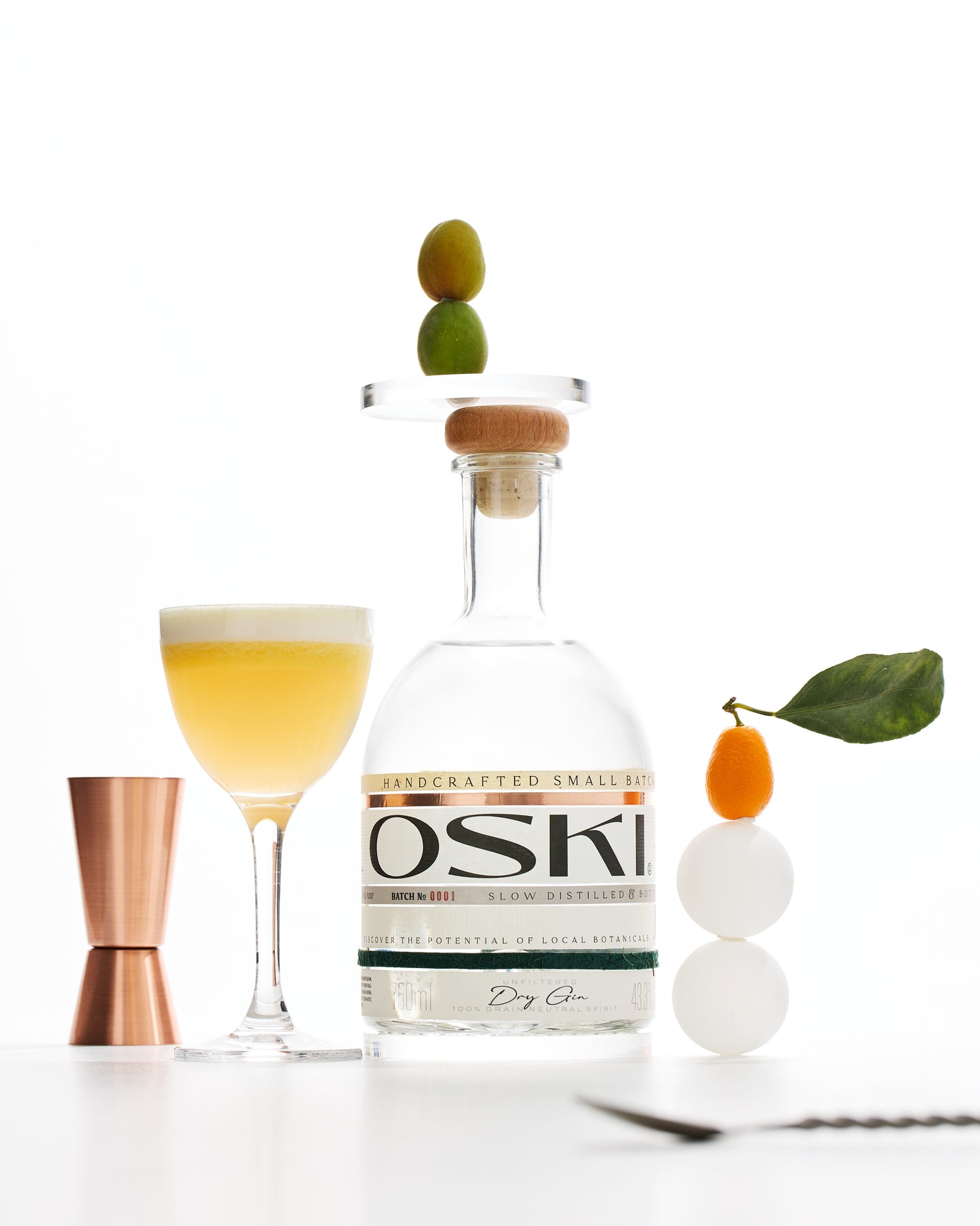 OSKI Gin bottle with a yellow cocktail in a coupe glass, garnished with citrus and styled alongside a copper jigger and fresh botanicals.