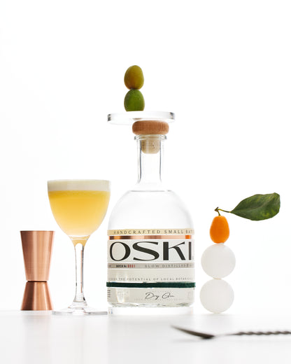 OSKI Gin bottle with a yellow cocktail in a coupe glass, garnished with citrus and styled alongside a copper jigger and fresh botanicals.