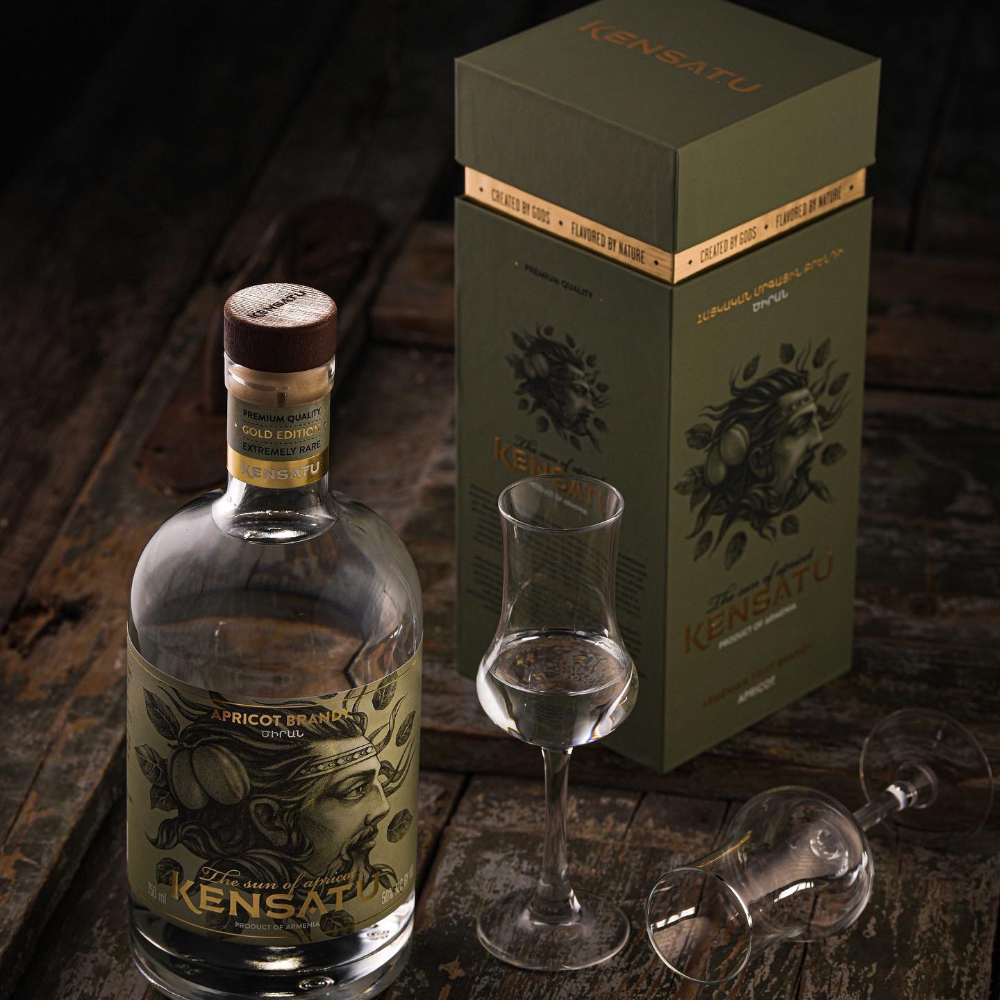 Kensatu Apricot Brandy bottle and gift box presented on a dark wooden surface with two elegant brandy glasses, one upright and one tipped over.